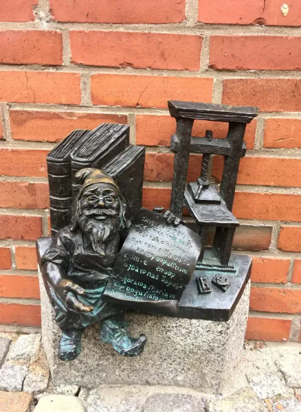 Bronze statue of a bespectacled dwarf holding a document, next to books and a printing press, against a red brick wall.