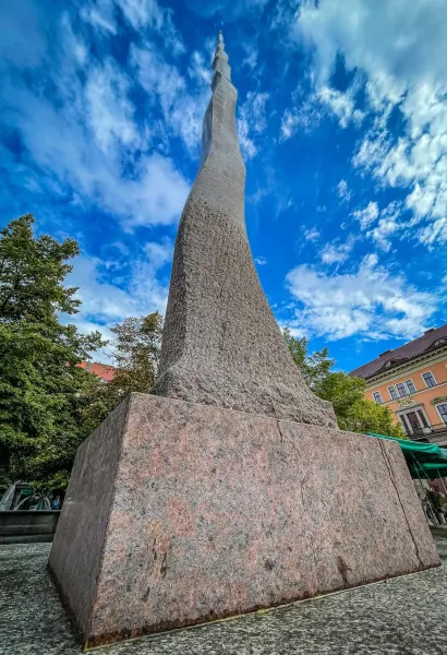 Wysoki, spiralny abstrakcyjny pomnik kamienny o chropowatej fakturze, na granitowej podstawie, pod błękitnym niebem z chmu...