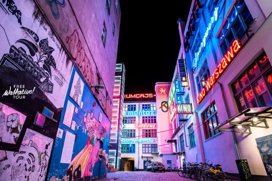 Wąska uliczka nocą z kolorowymi neonami na budynkach i ścianą pokrytą graffiti i muralami. Zaparkowane rowery.