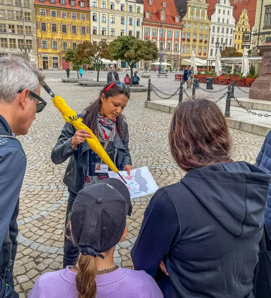 Przewodniczka z żółtym parasolem i mapą objaśnia trójce turystów na brukowanym placu. W tle kolorowe zabytkowe kamienice.