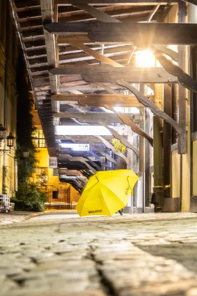 Żółty parasol Walkative! na brukowanej ulicy nocą, pod drewnianym pasażem oświetlonym jasnym światłem.