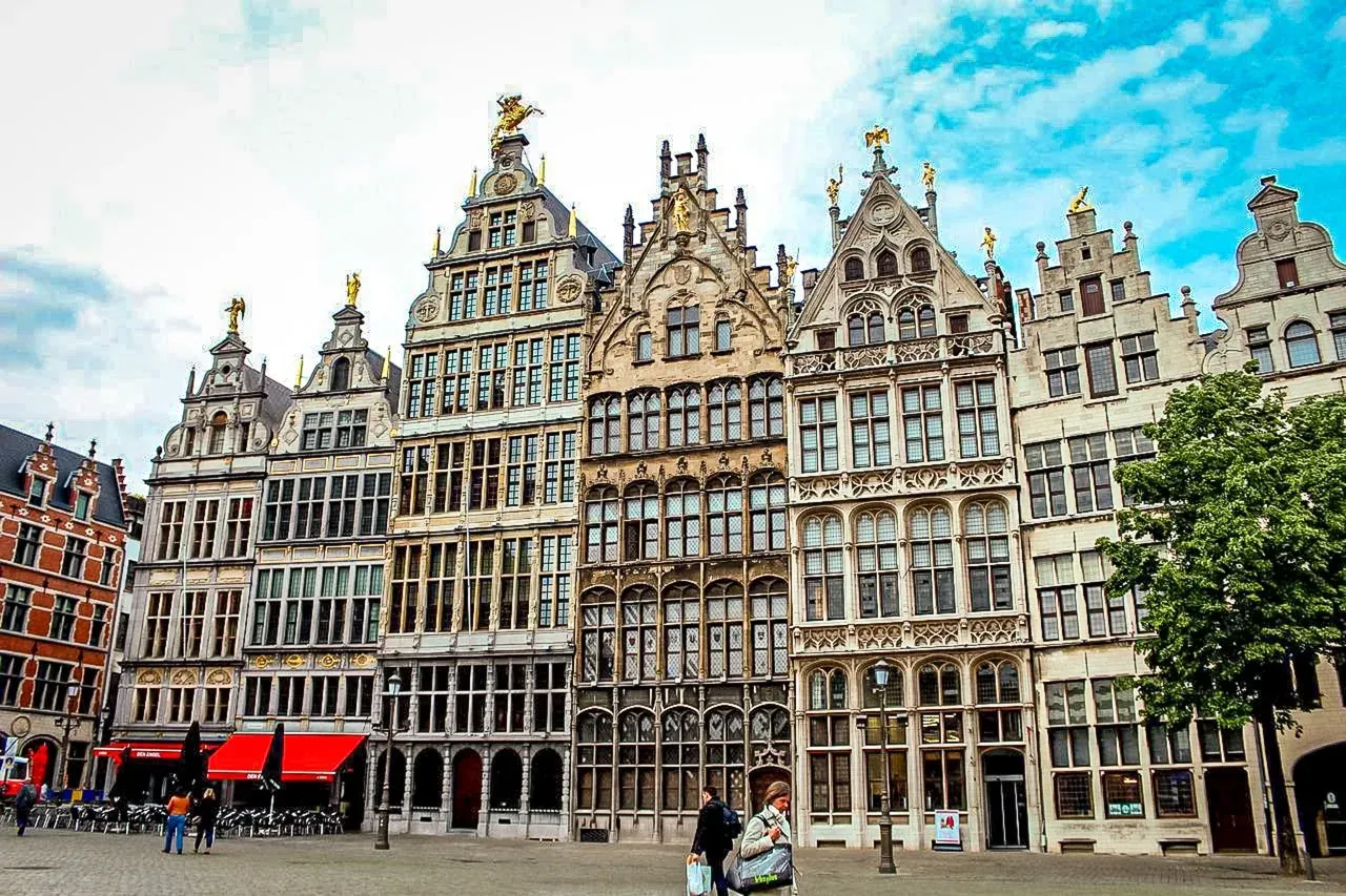 Grandes casas gremiales históricas con tejados escalonados y estatuas doradas en una plaza adoquinada de Amberes. Gente camin