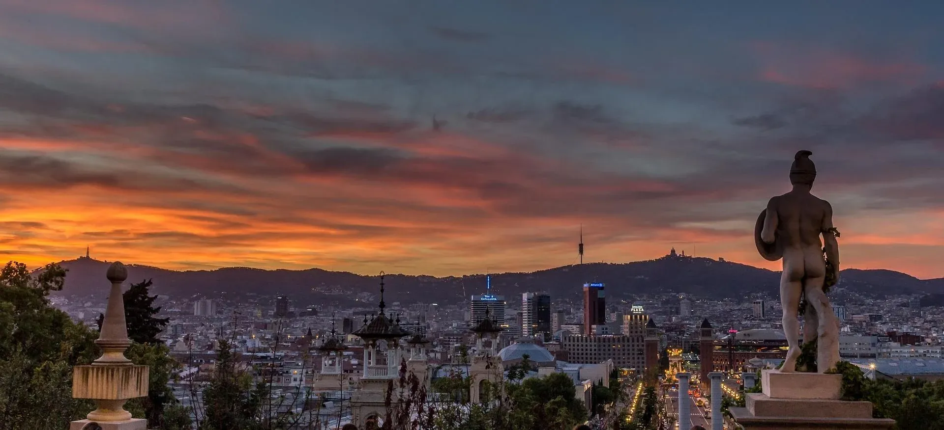 Panoramic view of Barcelona at sunset, with a statue in the foreground and Tibidabo Mountain in the distance.