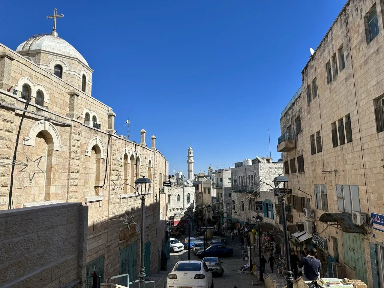 A narrow Bethlehem street with a church dome and cross on one side and a minaret in the distance.