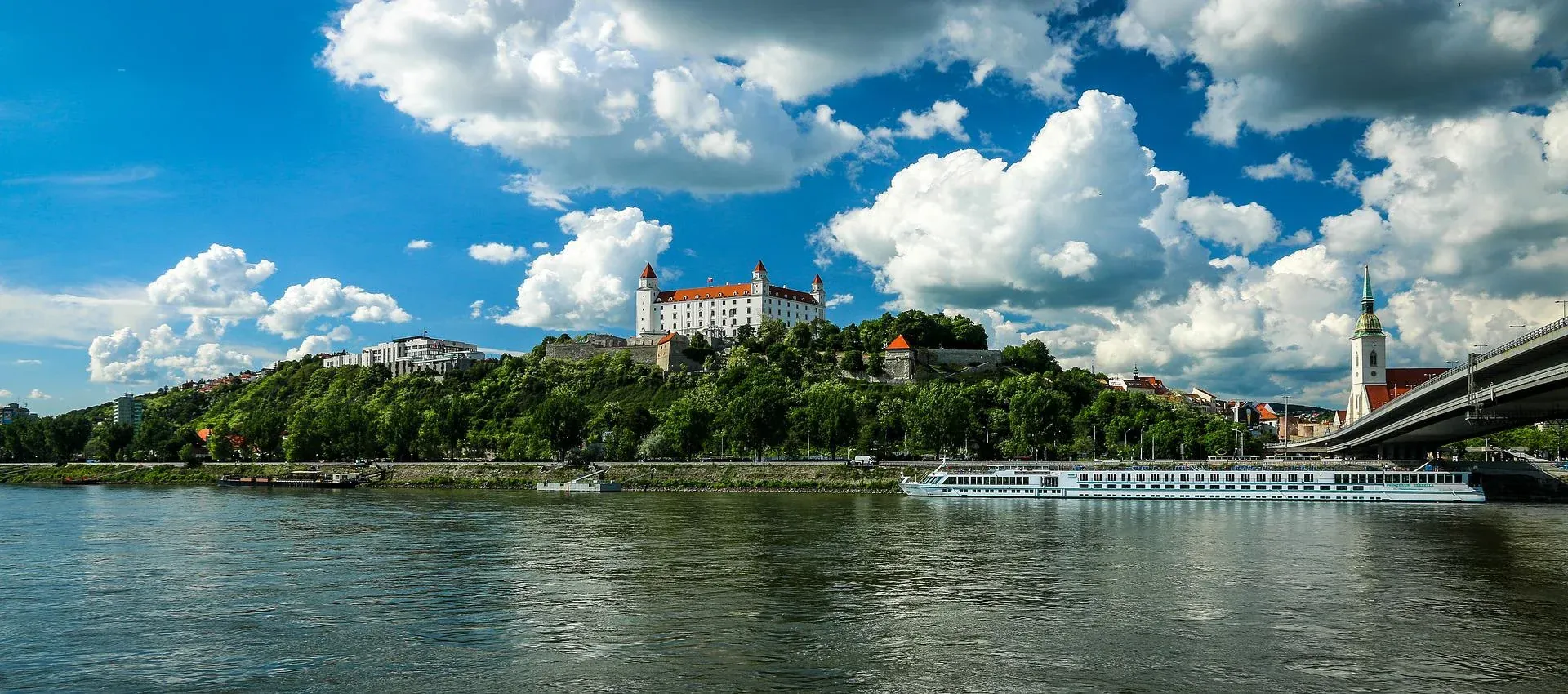 Castillo de Bratislava en una colina verde sobre el río Danubio, con un crucero, puente y aguja de la Catedral de San Martín.