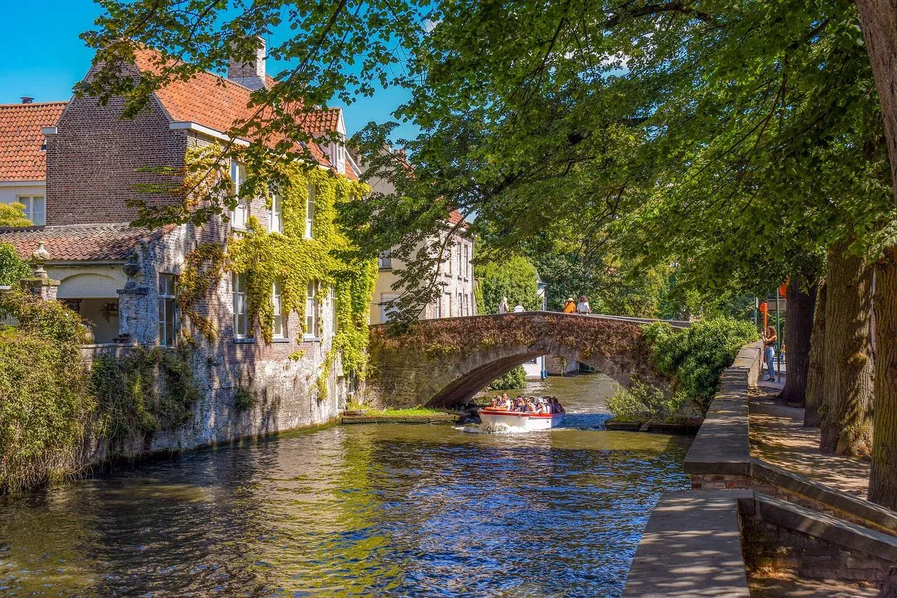 A tour boat with passengers on a canal in Bruges, passing under a stone arch bridge next to an ivy-covered building.