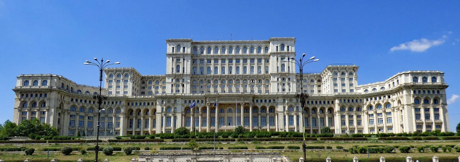 The Palace of the Parliament in Bucharest, a massive white building with many windows and columns, set against a blue sky wit