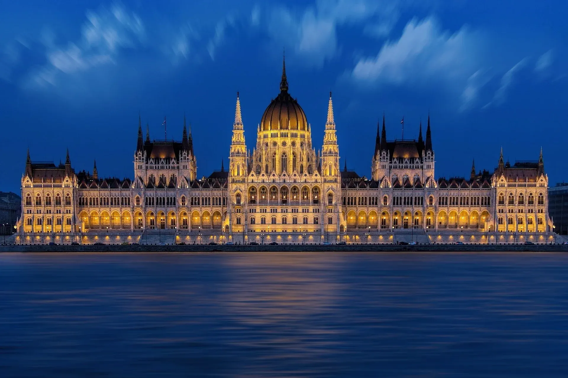 Illuminated Hungarian Parliament building at night, viewed from across the Danube River with blue sky.