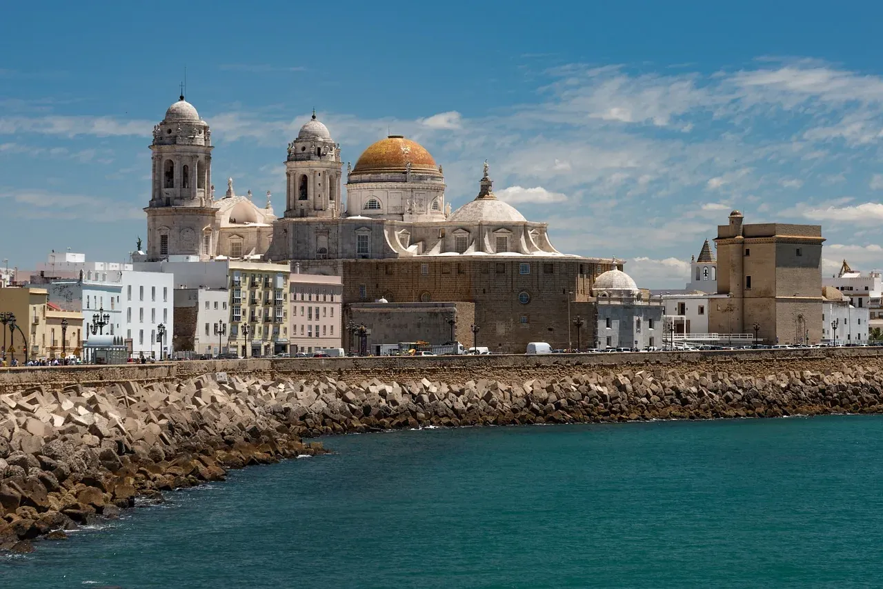 Blick auf die Kathedrale von Cádiz und die Skyline der Stadt vom Meer aus, mit einer steinigen Mole im Vordergrund unter blau
