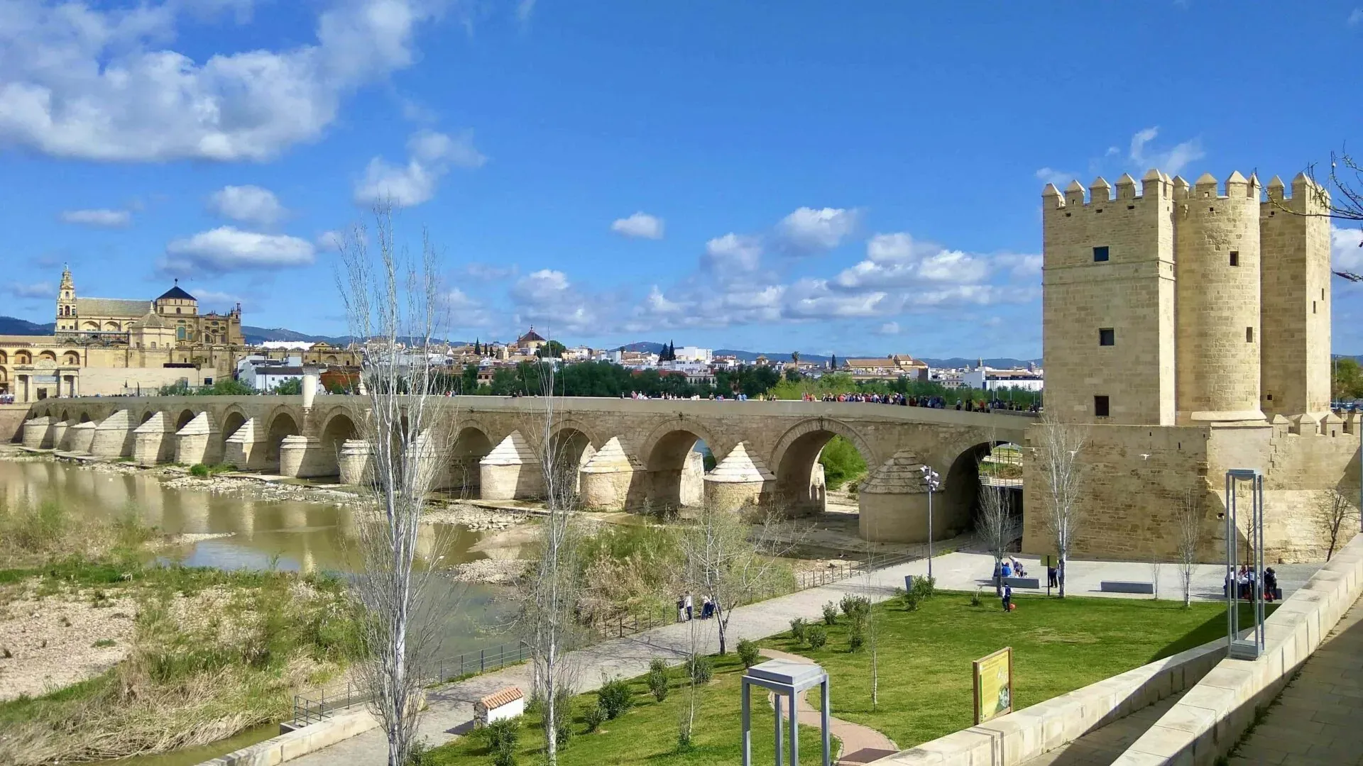 Puente Romano sobre el río Guadalquivir en Córdoba, con Torre de la Calahorra y Mezquita-Catedral al fondo bajo cielo azul.