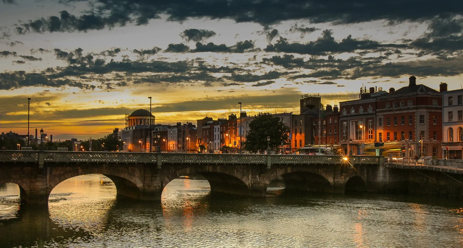 Dublin cityscape at sunset with Ha'penny Bridge over the River Liffey, glowing warm light, and historical buildings.