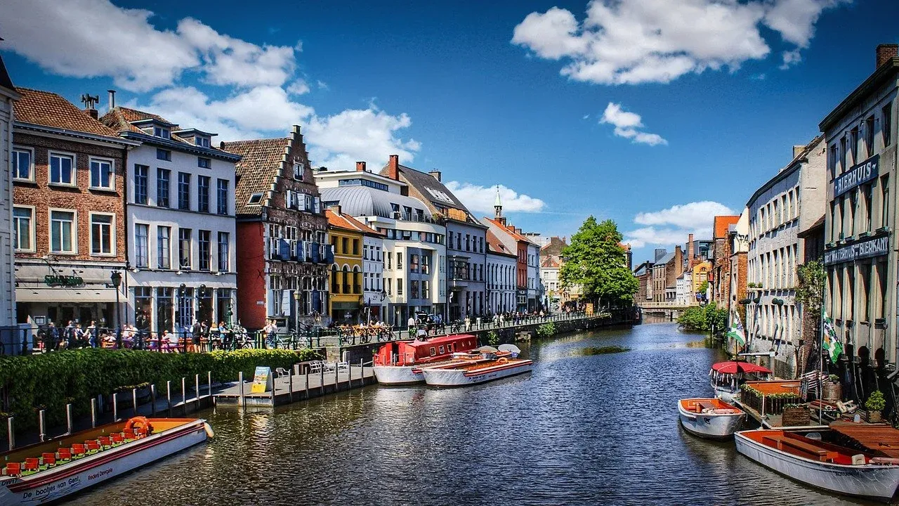 A sunny view of a canal in Ghent lined with historic, colorful buildings and several tour boats docked along the side.