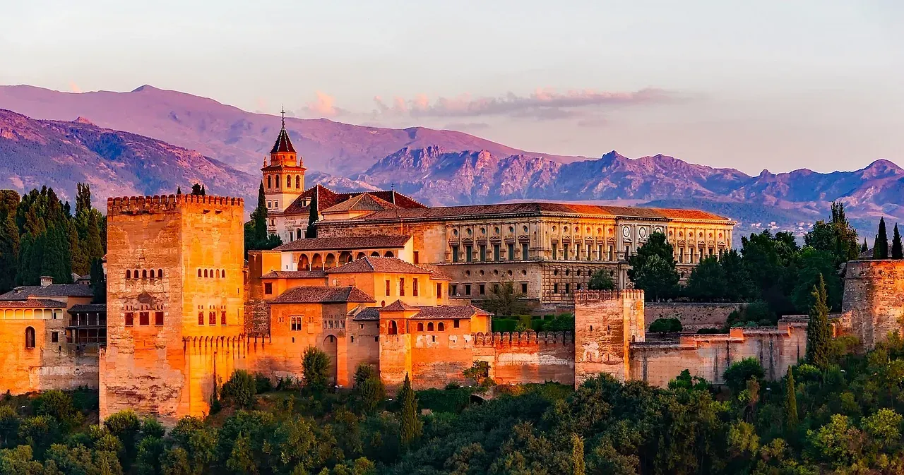 Alhambra-Palastkomplex mit Befestigungen und Türmen bei Sonnenuntergang, Berge im Hintergrund, umgeben von grünen Bäumen.