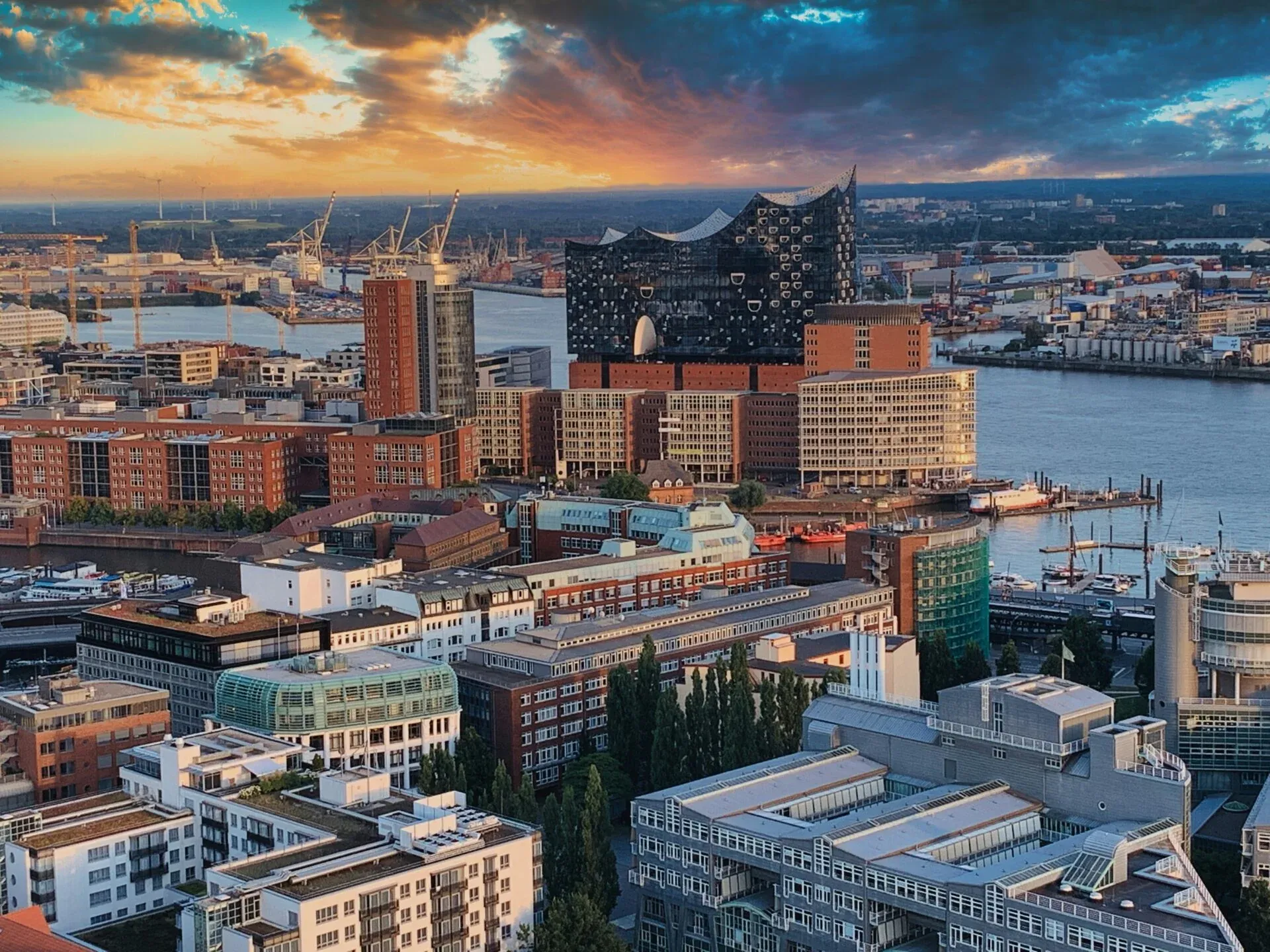 Aerial view of Hamburg harbor at sunset, featuring the Elbphilharmonie, brick buildings, and industrial cranes by the water.
