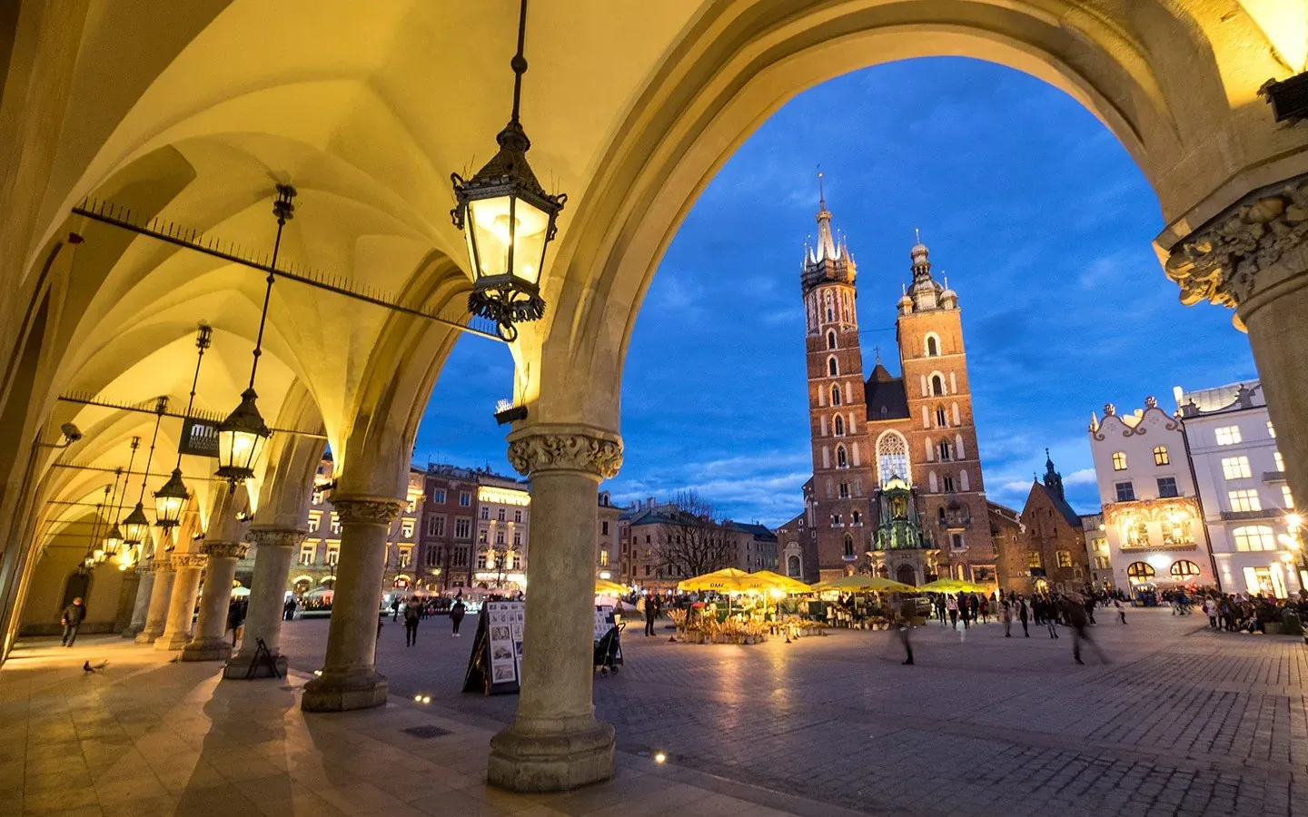 View from an illuminated archway of the Krakow Main Market Square with St. Mary's Basilica at dusk.
