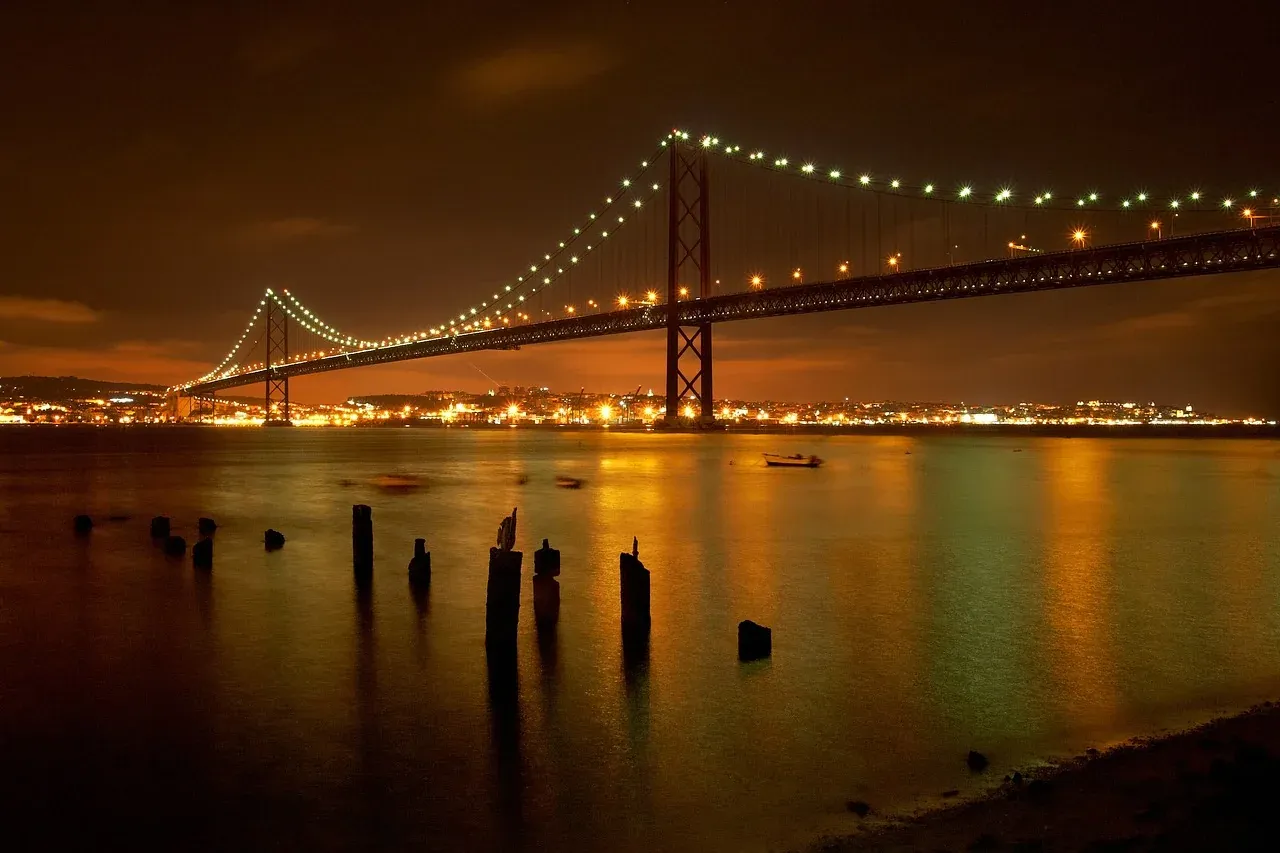 25 de Abril Bridge illuminated at night over the Tagus River, with city lights and reflections on the water.