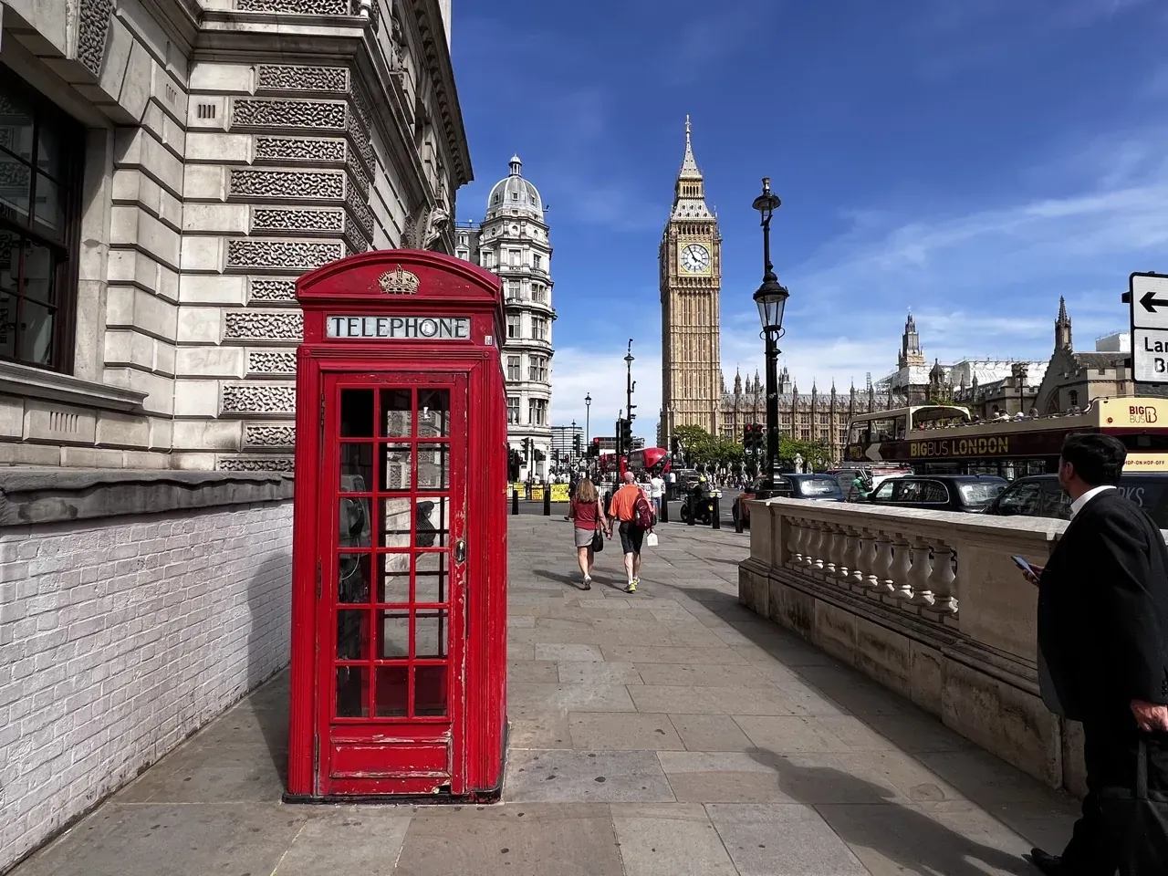 Klassische rote Telefonzelle auf einem Londoner Gehweg mit Big Ben und den Houses of Parliament im Hintergrund unter blauem H
