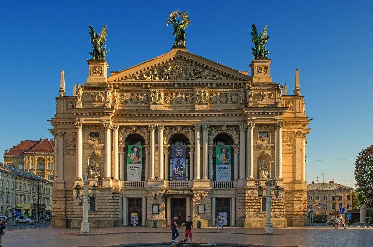 Facade of the Lviv Opera House with columns, sculptures on the roof, and banners. Clear blue sky above. People walk in the fo