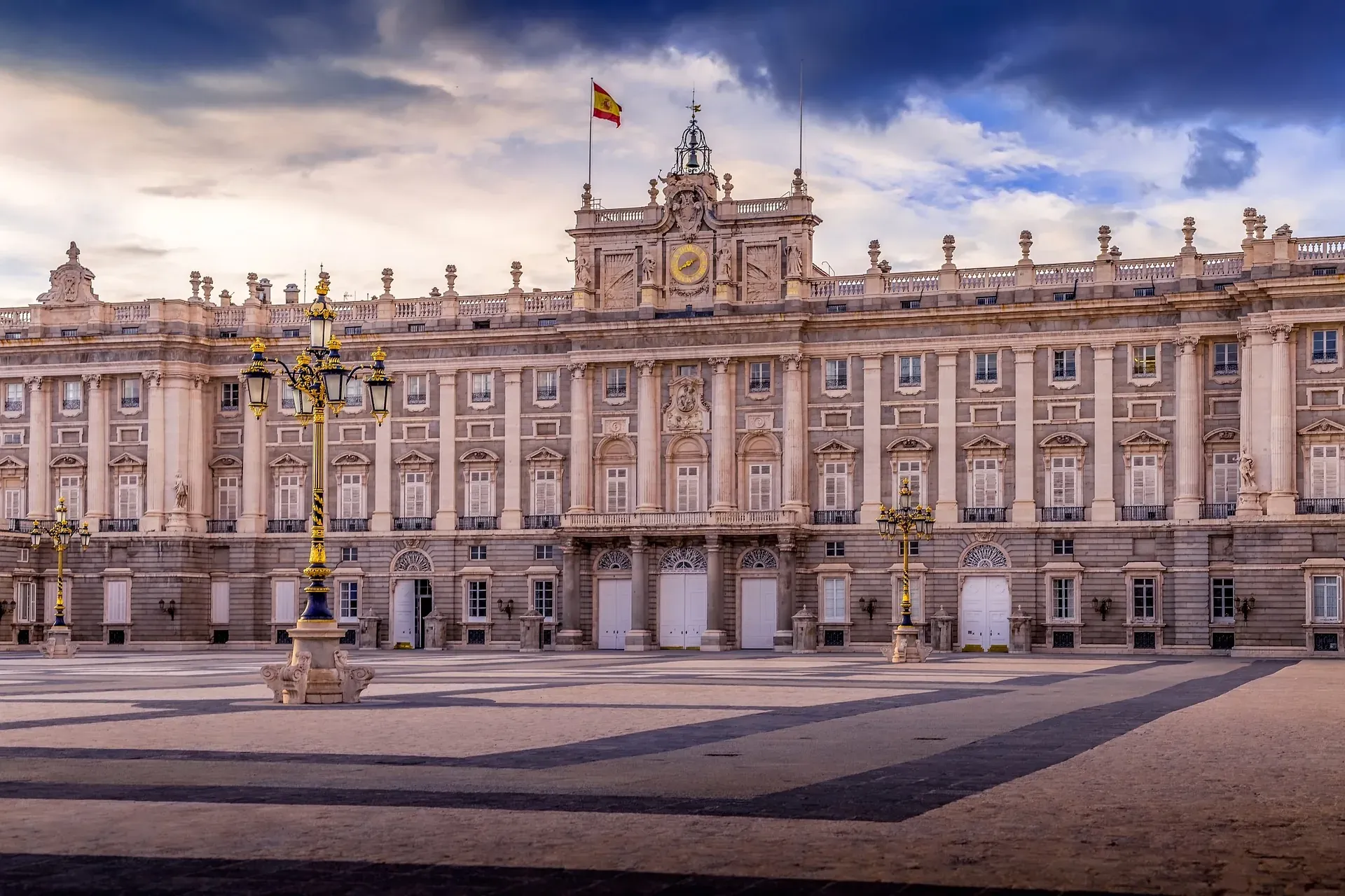 Exterior view of the Royal Palace of Madrid under a dramatic sky, with cobblestone plaza and lampposts.
