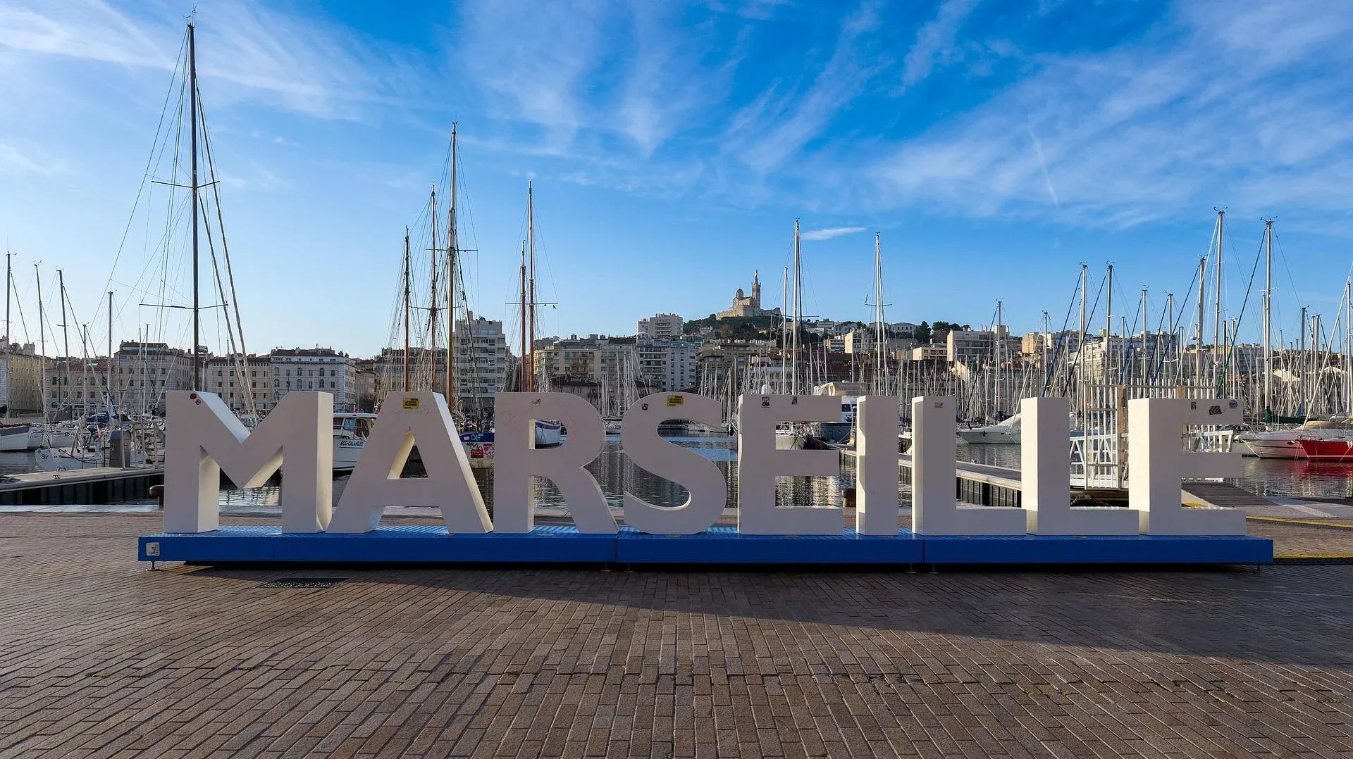 Grandes letras MARSEILLE sobre base azul en el Vieux-Port, con veleros y edificios. Notre-Dame de la Garde visible en la coli