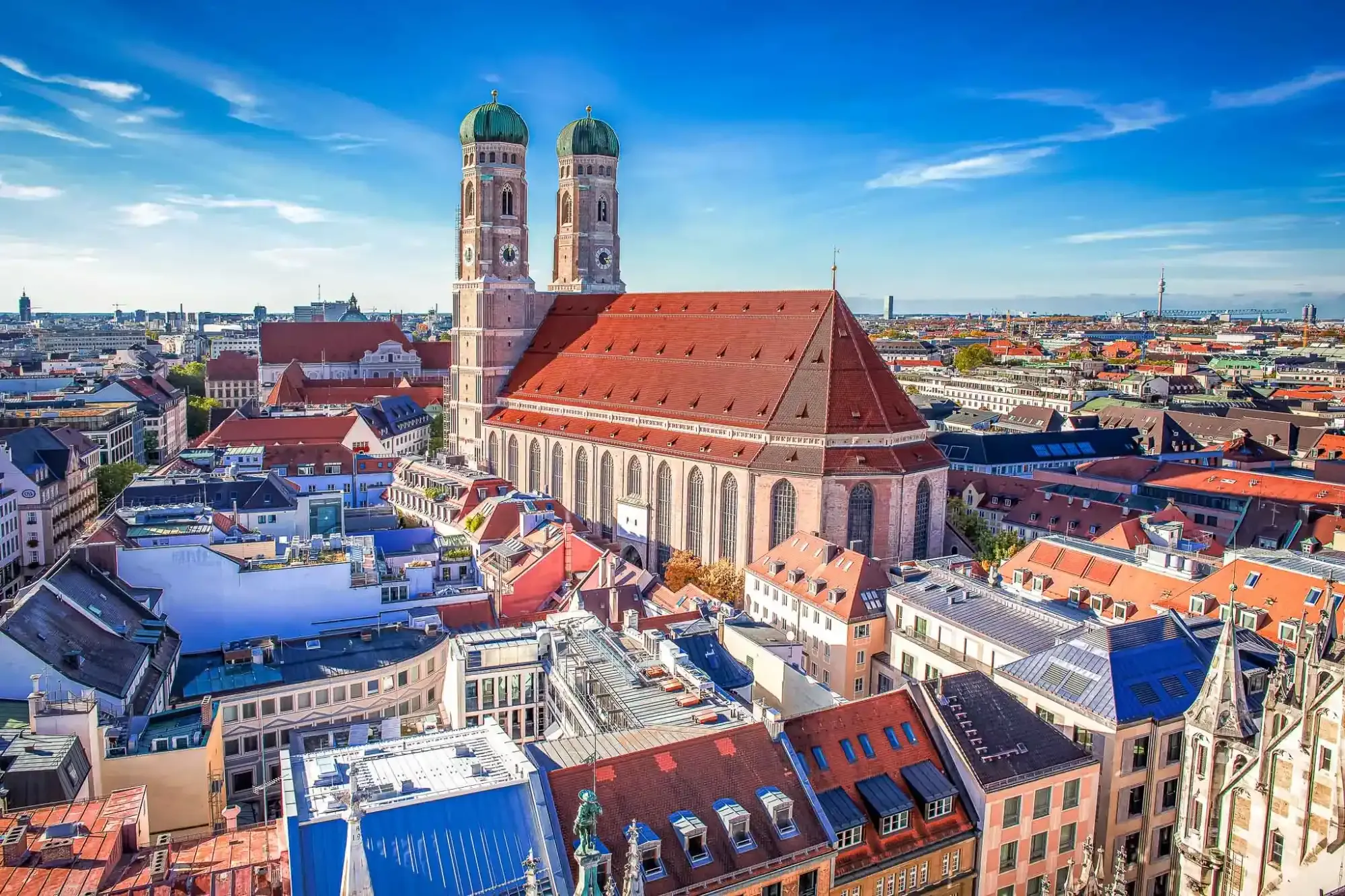 Luftaufnahme der Münchner Frauenkirche mit ihren zwei grünen Kuppeltürmen, umgeben von Stadtdächern.