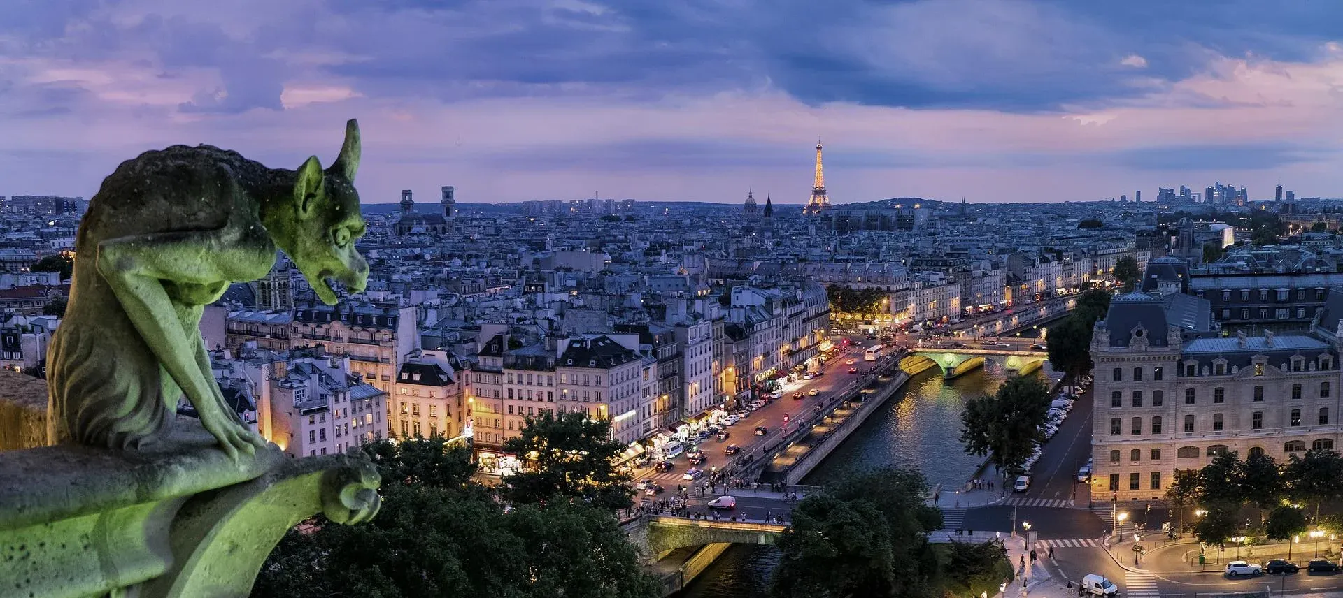 Stone gargoyle on a building overlooking the Paris cityscape at dusk, with the illuminated Eiffel Tower and city lights.