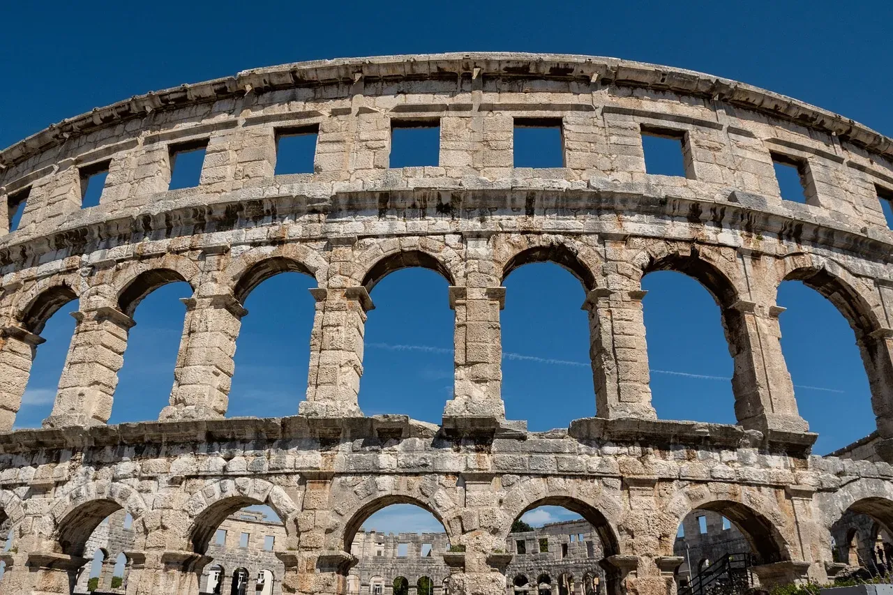 Exterior of the Pula Arena, a Roman amphitheater, with its preserved stone arches against a clear blue sky.
