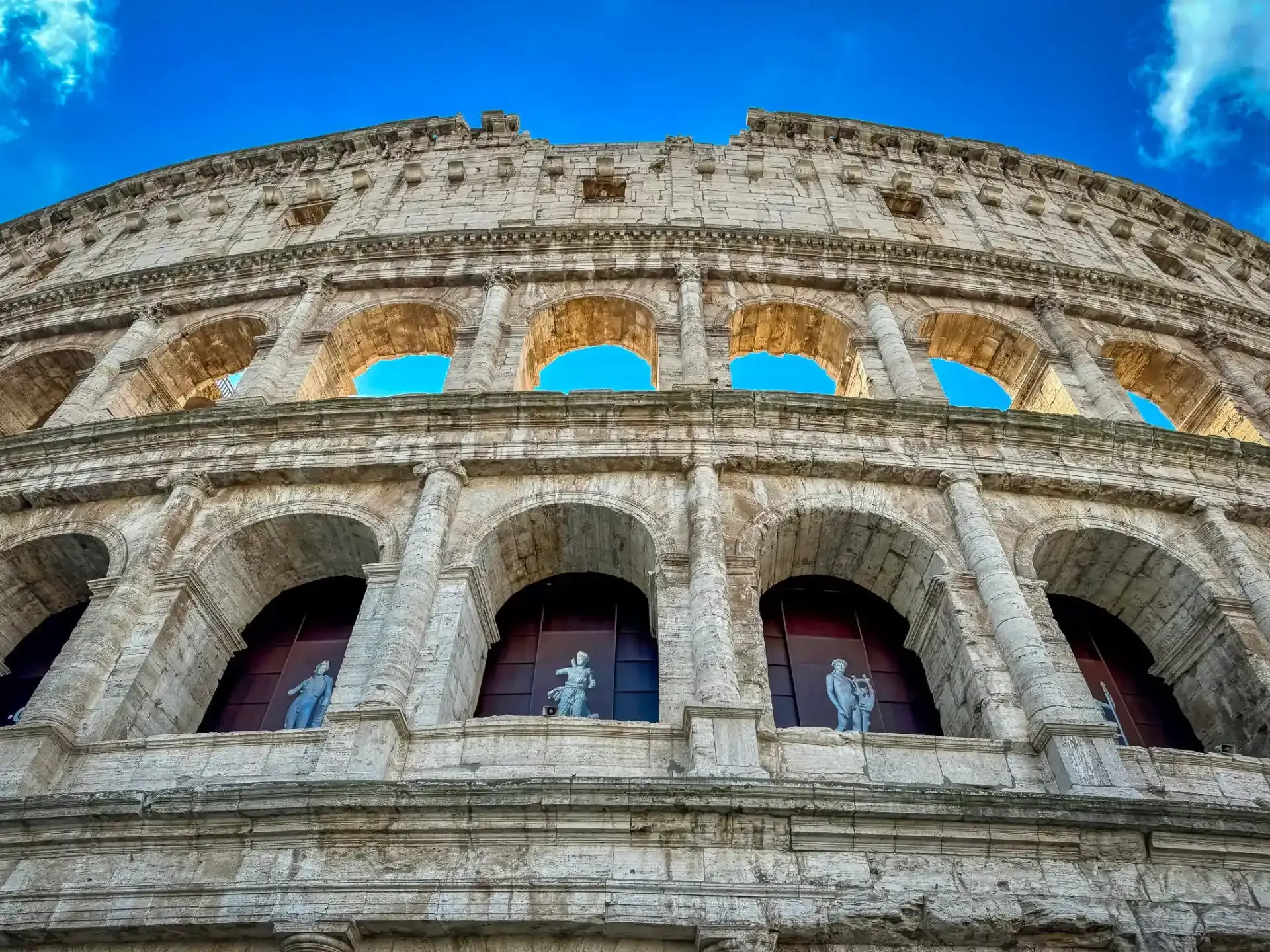 Zewnętrzny widok Koloseum w Rzymie, ukazujący kamienne łuki, kolumny i posągi w niższych arkadach, na tle błękitnego nieba.