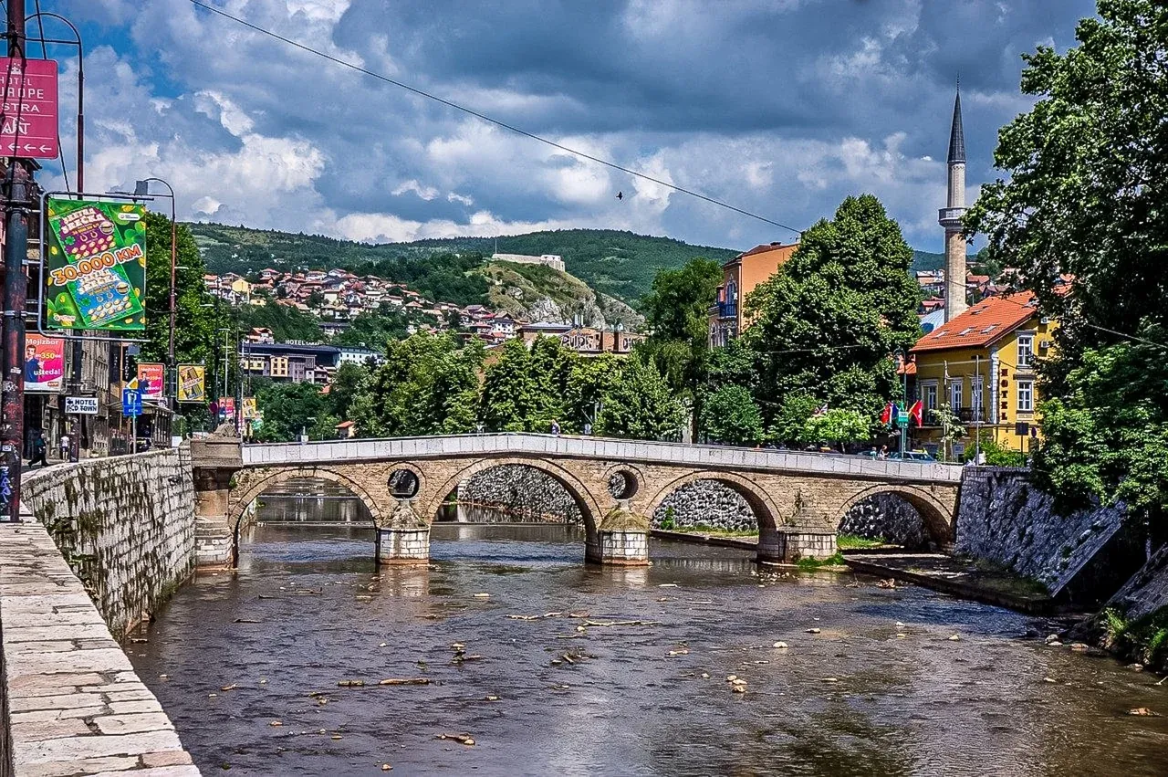 Historische Steinbogenbrücke über einen Fluss in Sarajevo, mit einem Minarett, Gebäuden und grünen Hügeln unter bewölktem Him