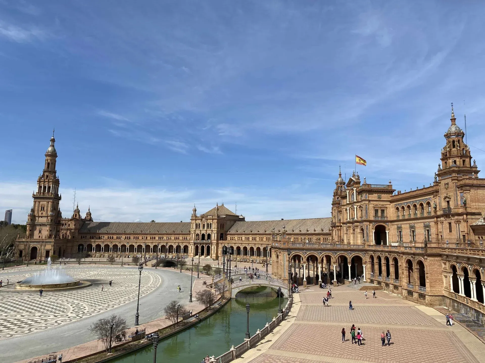 Large ornate building with arcades, a central canal with bridges, patterned plaza, and fountain under a blue sky in Seville.