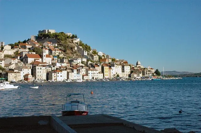 Sibenik old town and hilltop fortress from the sea, showing historic buildings with red roofs, boats, and a clear blue sky.