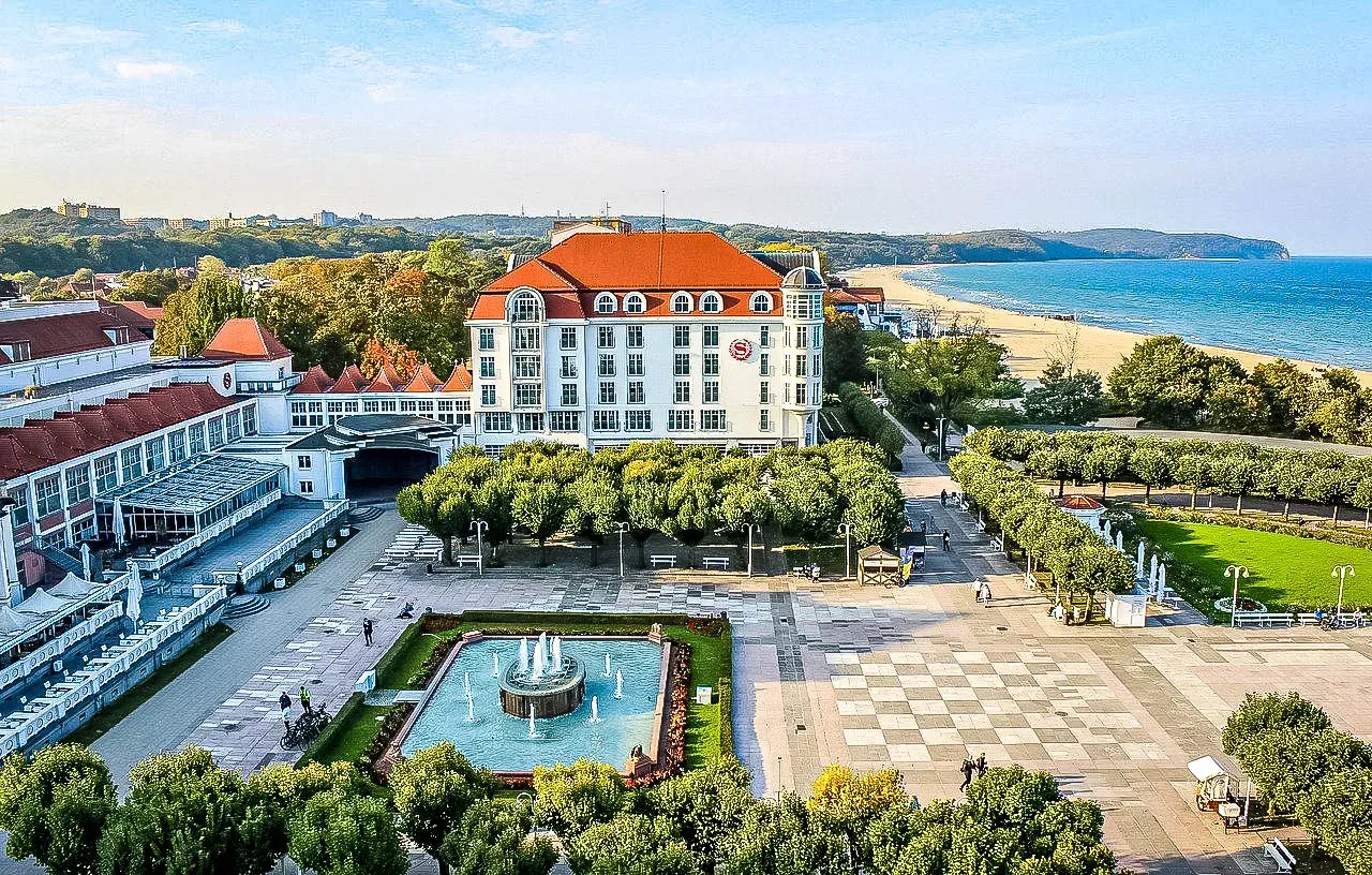 Luftaufnahme des Grand Hotels Sopot mit Platz, Brunnen, Bäumen, Sandstrand und Ostsee unter blauem Himmel.