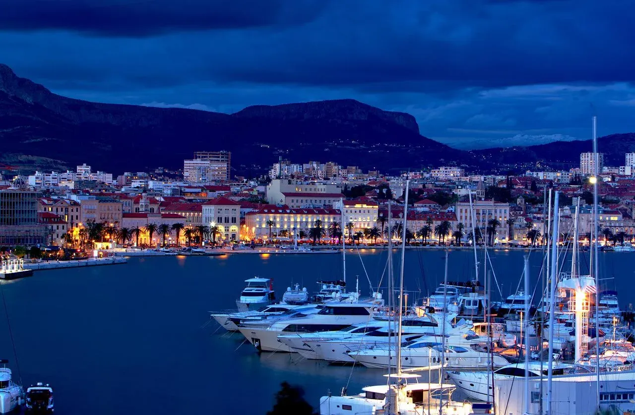 Split harbor at night, with illuminated city buildings, palm trees, mountains, and many yachts.