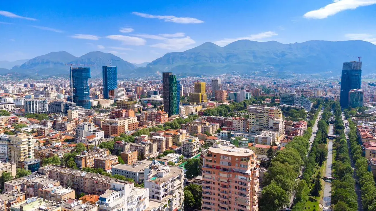 Luftaufnahme von Tirana mit modernen Hochhäusern, Wohngebieten, einer von Bäumen gesäumten Allee und Bergen im Hintergrund.
