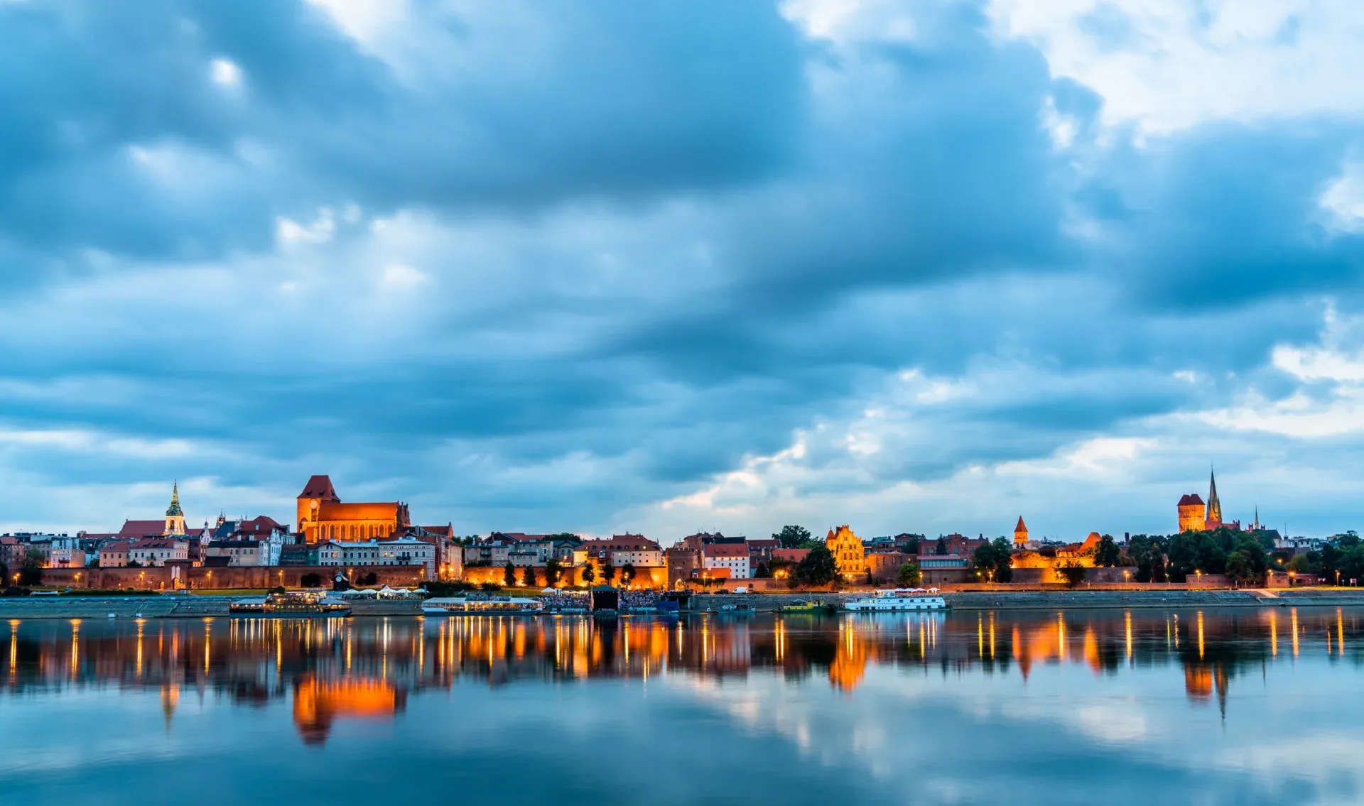 Toruń skyline at dusk, with historical buildings illuminated along the riverbank, reflected in the calm water under a cloudy