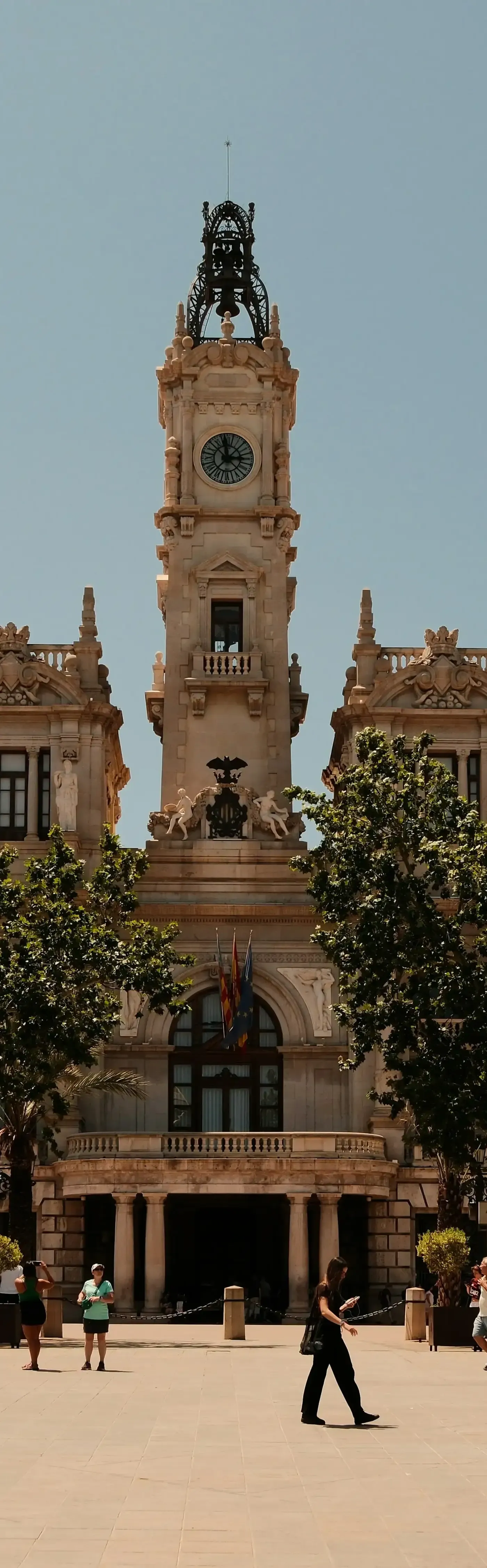 Torre del reloj ornamentada del Ayuntamiento de Valencia con gran esfera de reloj, estatuas, banderas y árboles verdes en pri