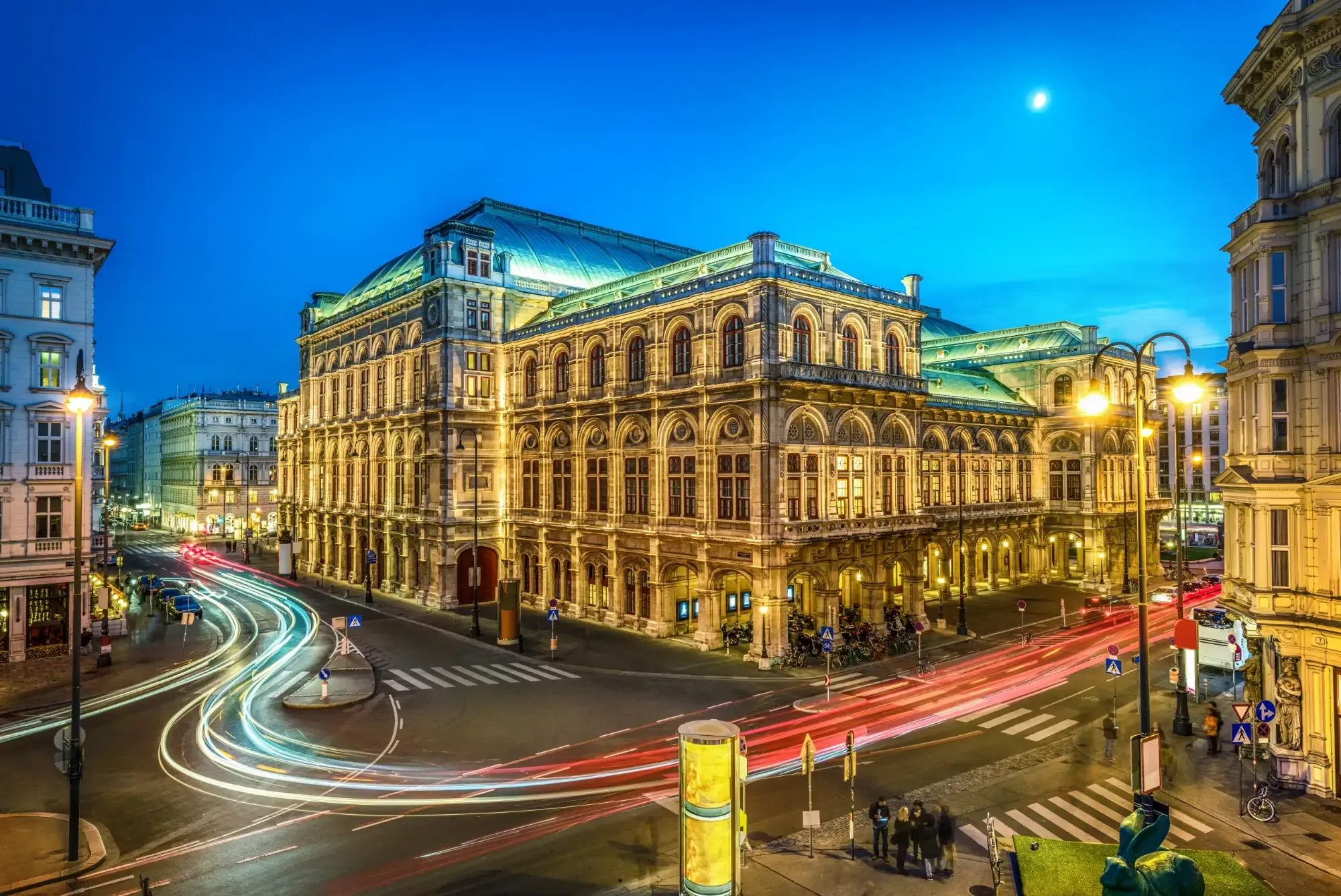 Beleuchtetes Wiener Staatsoperngebäude in der Dämmerung, mit Lichtspuren von Autos an einer Kreuzung und einem Halbmond am Hi