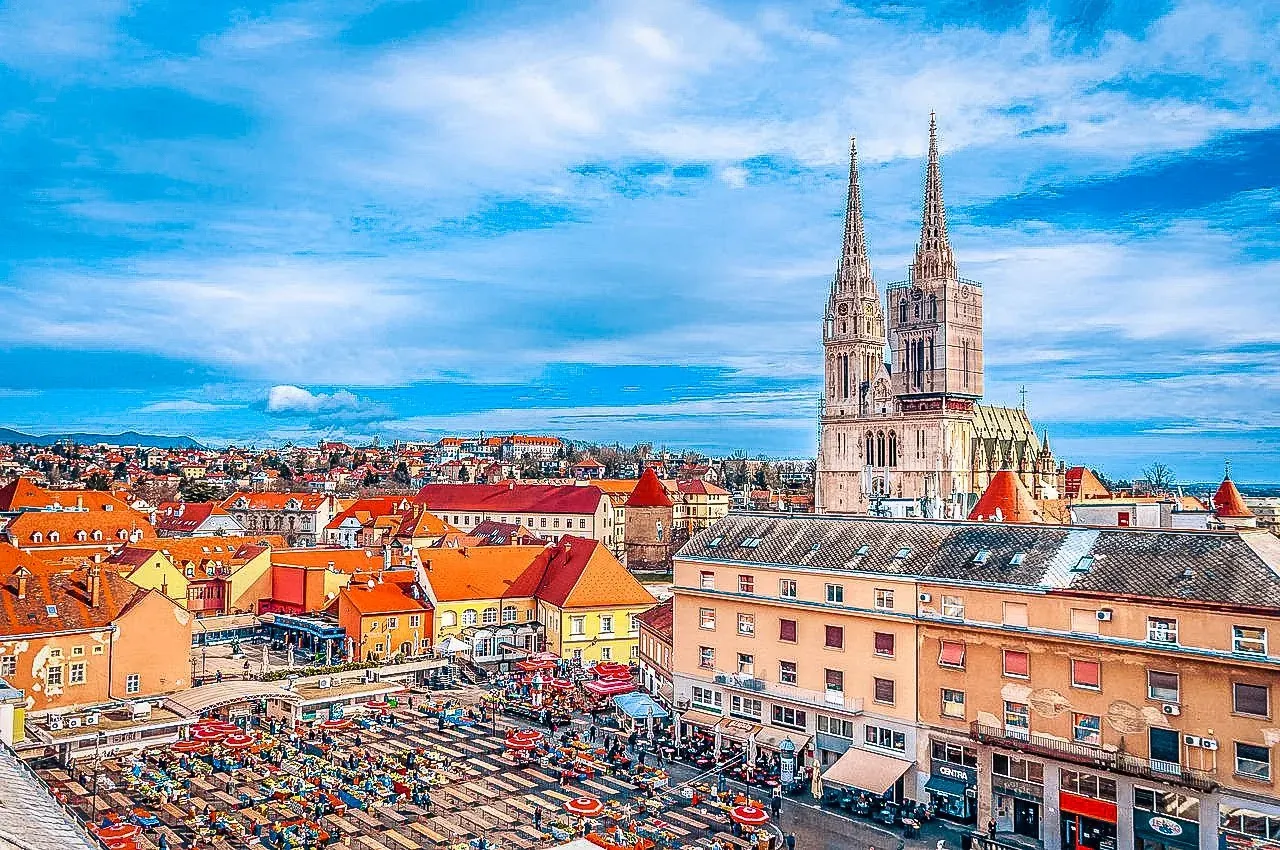Aerial view of Zagreb with the Cathedral featuring two spires, partially scaffolded, and a bustling outdoor market with red u