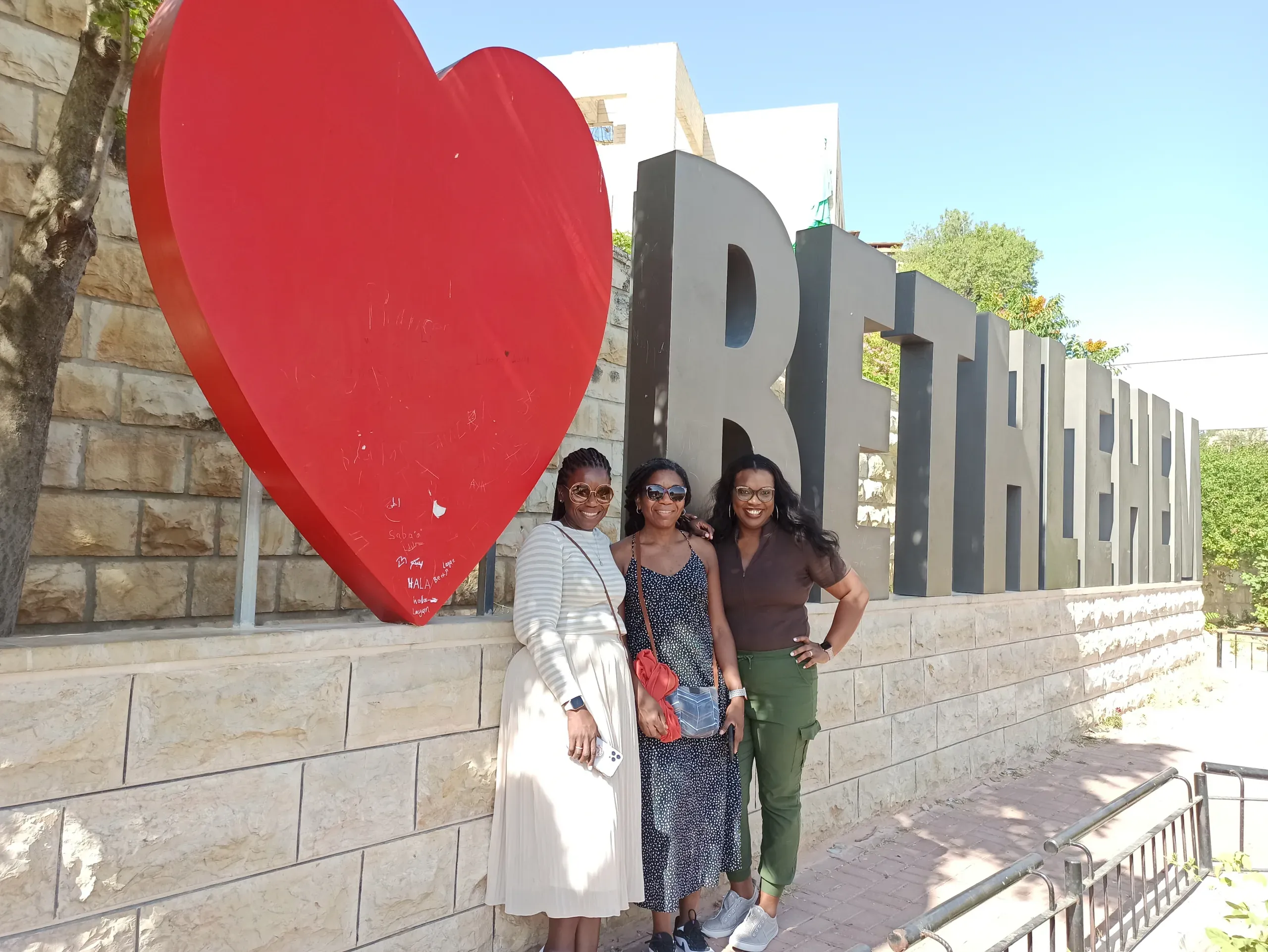 Three women pose happily in Bethlehem, Palestine, in front of a large red heart and the city's name.