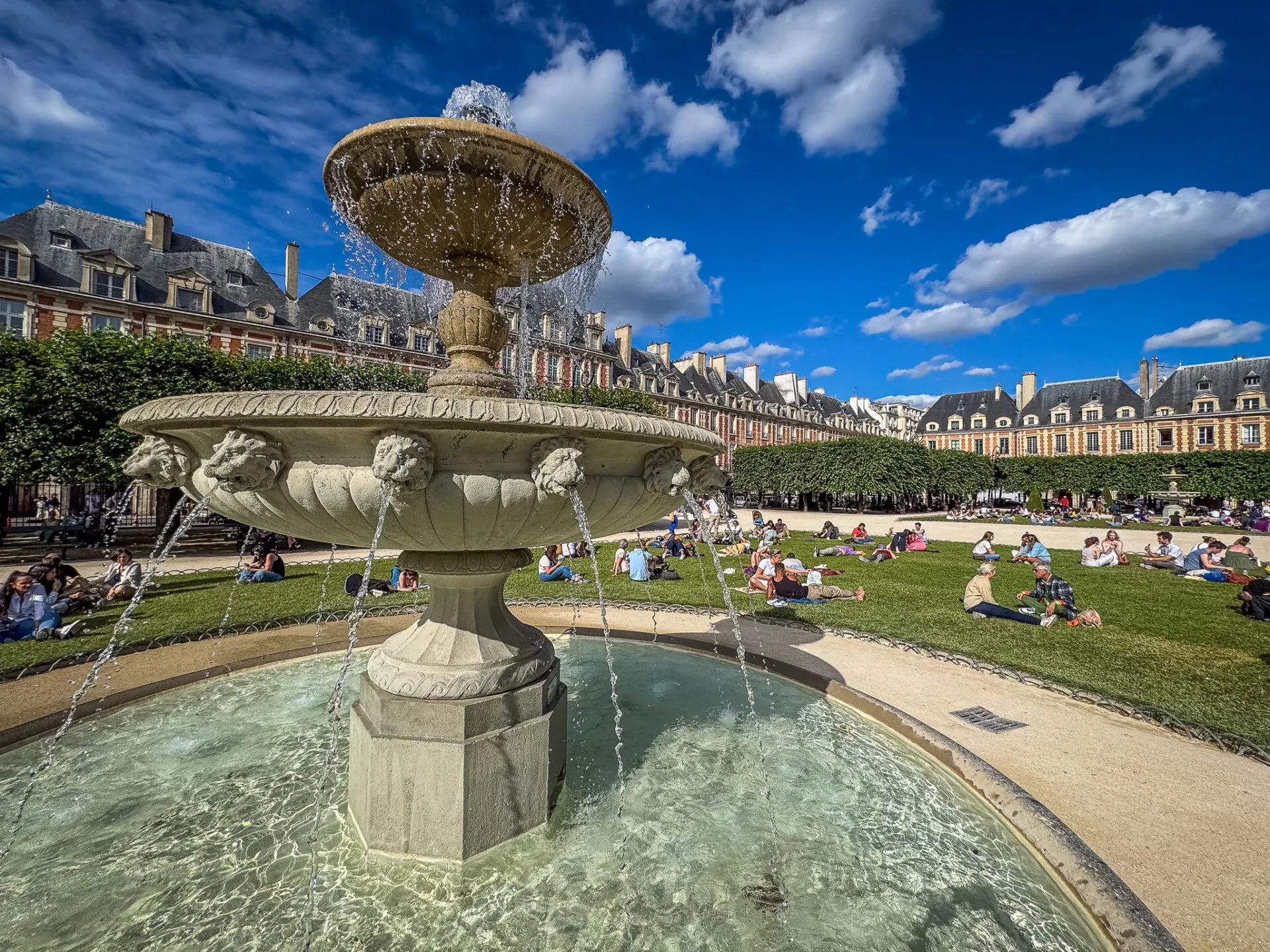 A picturesque fountain in Place des Vosges, Le Marais, Paris, surrounded by people enjoying a sunny day.