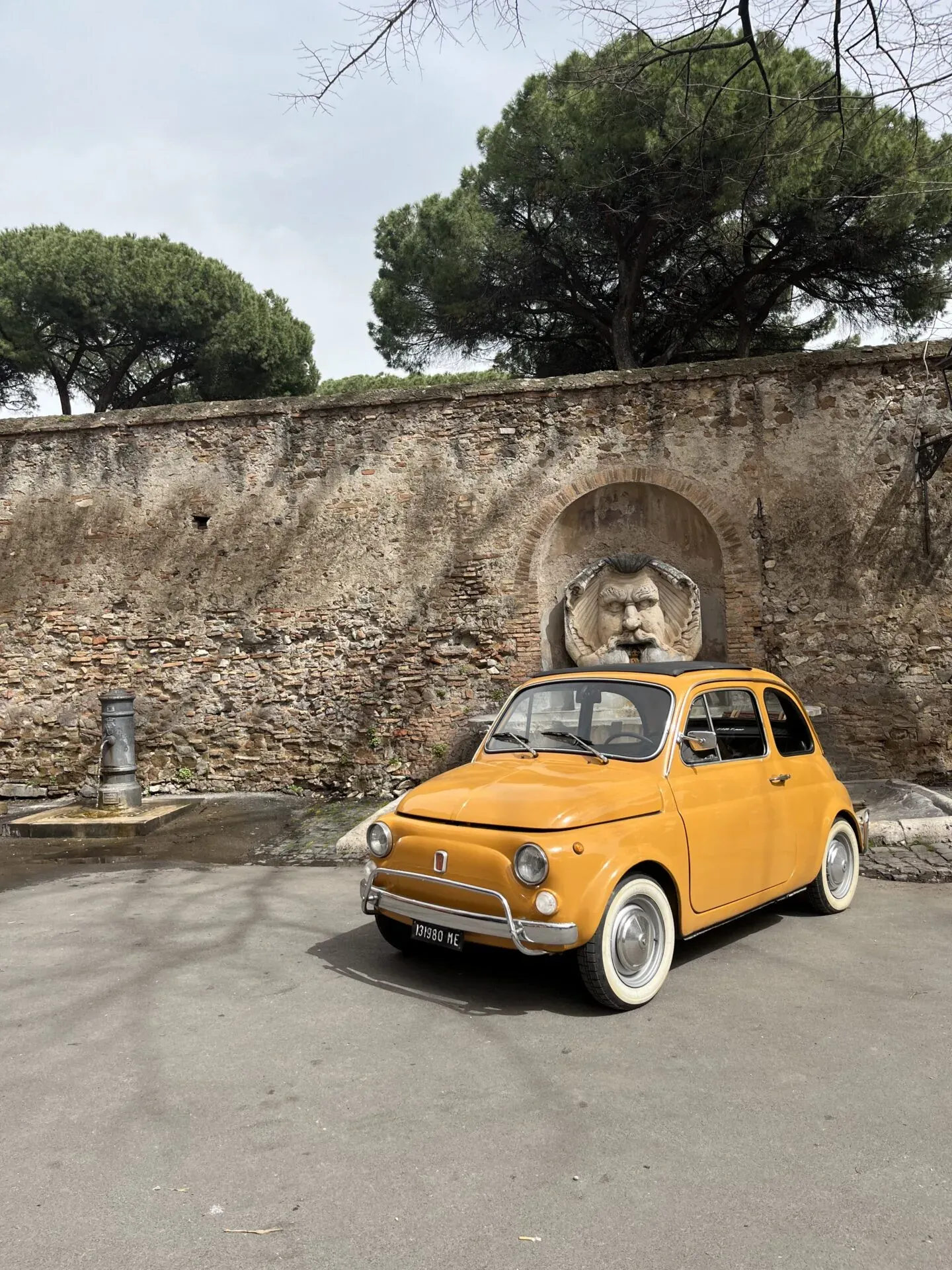 Ein gelber Fiat 500, geparkt an einer römischen Mauer mit einem einzigartigen Brunnen.