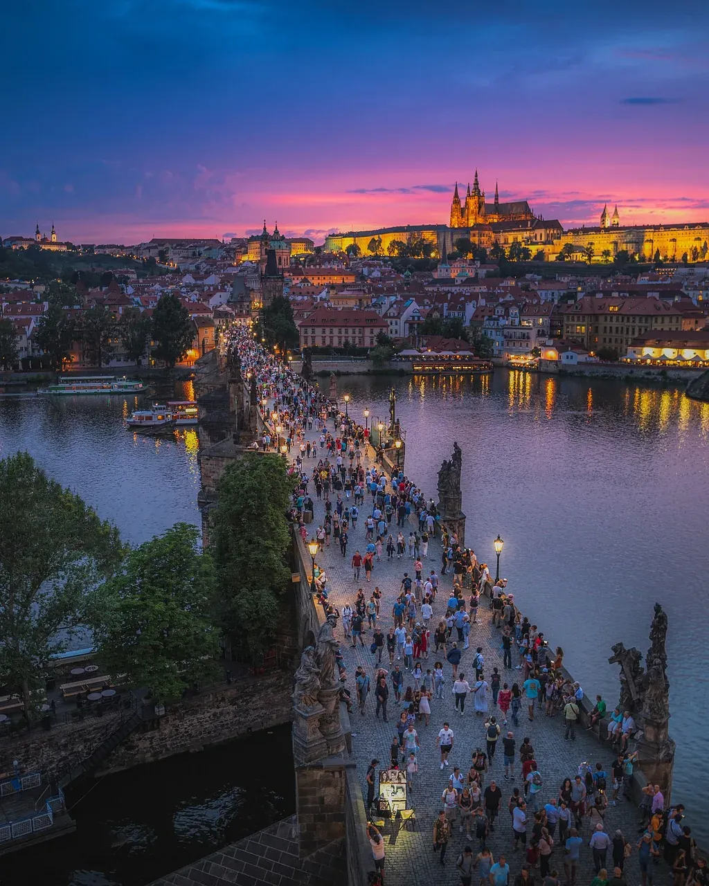 Turistas pasean por el Puente de Carlos en Praga al atardecer, con el Castillo de Praga majestuoso iluminado al fondo.