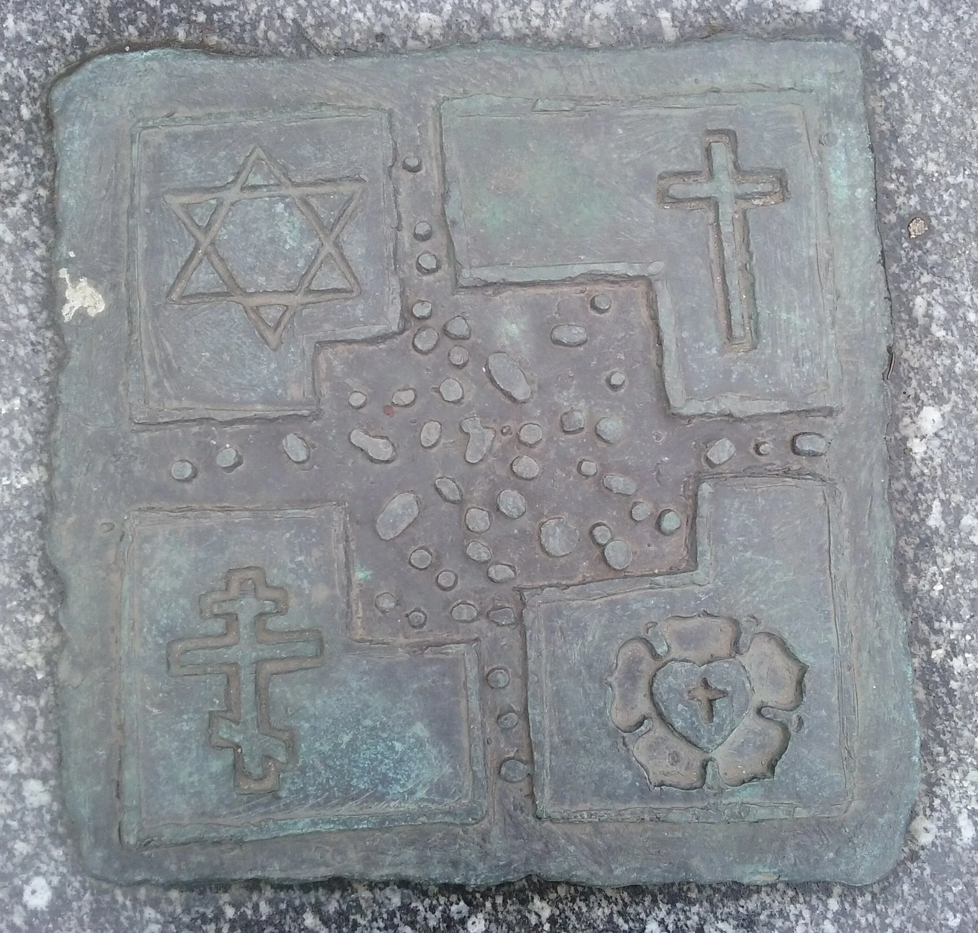 Bronze plaque depicting four religious symbols: Star of David, Christian cross, Orthodox cross, and a heart with a cross.